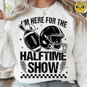 May include: White sweatshirt with a black graphic of a football, helmet, and the text "I'M HERE FOR THE HALFTIME SHOW." The design includes stars and checkered flag lightning bolts.