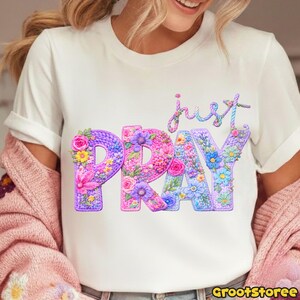 May include: White t-shirt with the words "just PRAY" in a floral design. The word "PRAY" is in large, colorful letters filled with flowers. The word "just" is in cursive above the word "PRAY".