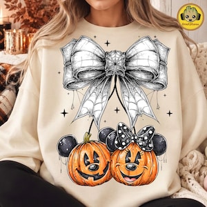 May include: A cream-colored sweatshirt featuring a large black and white graphic of a bow with a spiderweb pattern. Below the bow are two orange pumpkins with black mouse ears, one wearing a black and white polka dot bow, surrounded by stars.