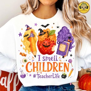 May include: A white sweatshirt featuring a graphic print of a yellow pencil with blonde hair, a red apple with a jack-o'-lantern face and orange hair, and a purple book with purple hair. Text reads I smell CHILDREN #TeacherLife.