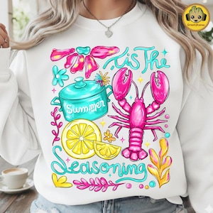 Puede incluir: Sudadera blanca con un diseño gráfico colorido. El diseño incluye una langosta rosa, una olla turquesa con la palabra "Summer" y una rodaja de limón. También aparece el texto "'Tis The Seasoning".