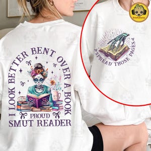 May include: White t-shirt with a purple and white design of a skeleton woman reading a book. The text on the shirt reads "I look better bent over a book, proud smut reader".