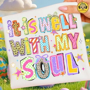 May include: White t-shirt with the phrase "it is well with my soul" in colorful, block letters. The letters feature various patterns, including leopard print and checkered designs, with star and bow accents. The shirt is held by a hand with a pink nail.