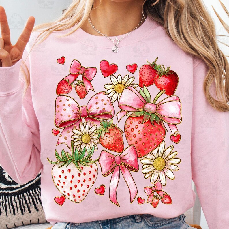 May include: Pink sweatshirt with a graphic design of strawberries, bows, hearts, and daisies. The design is in a watercolor style with gold accents.