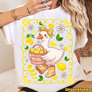 May include: White t-shirt featuring a cartoon goose with a yellow bow, holding a basket of lemons. The design includes lemons, daisies, and green leaves, framed by a yellow border. The goose wears yellow rain boots with floral patterns.