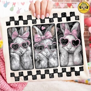 May include: A decorative pillow featuring three grayscale illustrations of rabbits. Each rabbit wears pink heart-shaped sunglasses and a pink bow. The pillow has a black and white checkered border.
