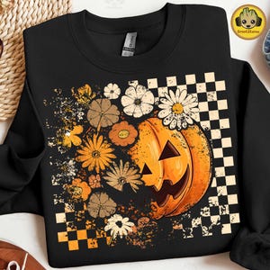 Op de afbeelding: Zwarte sweatshirt met een Halloween-ontwerp. Het heeft een oranje pompoen met een uitgesneden gezicht, omringd door verschillende bloemen in tinten oranje, bruin en wit. Een dambordpatroon is te zien aan de rechterkant van de pompoen.