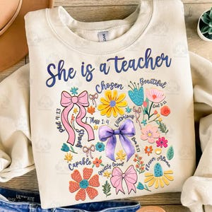 Puede incluir: Una sudadera beige con el texto "She is a teacher" en letras azules. El dise&ntilde;o presenta una forma de coraz&oacute;n hecha de flores, lazos y vers&iacute;culos b&iacute;blicos.