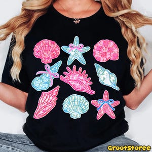 May include: Black t-shirt featuring a collection of colorful seashell and starfish illustrations. The designs include pink, blue, and white shells and starfish, some adorned with bows. The shirt has a round neckline.