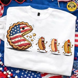 May include: White crew neck sweatshirt featuring a cartoon pie with the American flag design eating three running hot dogs. The pie has a face and is mid-bite, with crumbs falling. The hot dogs have panicked expressions.