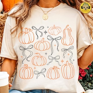 May include: A cream-coloured t-shirt featuring a repeating graphic print of orange line-art pumpkins and black line-art bows interspersed with small four-pointed star shapes across the front of the garment.