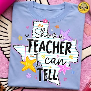 以下が含まれることがあります： ライトブルーのクルーネックTシャツ。罫線入りのノート用紙柄のテキサス州の形をしたグラフィックがプリントされています。黒い文字で「She's a TEACHER I can TELL」と書かれており、カラフルな星や小さな花のイラストが周囲に描かれています。