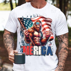 May include: White t-shirt featuring a cartoon illustration of a Bigfoot character wearing American flag sunglasses and holding a flag. The word "'MERICA" is printed in red, white, and blue. The shirt is worn by a person.