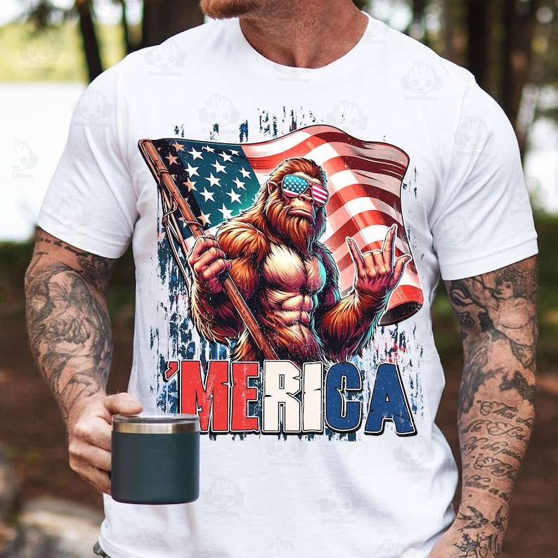 May include: White t-shirt featuring a cartoon illustration of a Bigfoot character wearing American flag sunglasses and holding a flag. The word "'MERICA" is printed in red, white, and blue. The shirt is worn by a person.