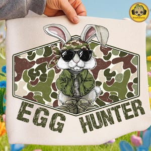 May include: A light-coloured fabric item with a cartoon rabbit wearing sunglasses, a camouflage bandana, and a green jacket. The rabbit is set against a camouflage pattern within a hexagonal frame. The text "EGG HUNTER" is printed below the rabbit in a distressed font.