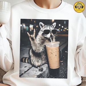 May include: White sweatshirt featuring a graphic of a raccoon wearing sunglasses and holding a coffee drink. The raccoon is making a rock and roll hand gesture. The background is a blurred city scene.