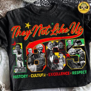 Peut inclure: T-shirt gris foncé avec l'inscription "They Not Like Us 1865" en rouge et vert. Le chiffre 1865 est rempli d'images de personnalités historiques. Les mots "History, Culture, Excellence, Respect" sont imprimés en bas.