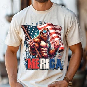 May include: Cream-colored t-shirt featuring a graphic of a muscular Bigfoot wearing American flag sunglasses and holding a flag. The flag is the American flag. The word "'MERICA" is printed in red, white, and blue.