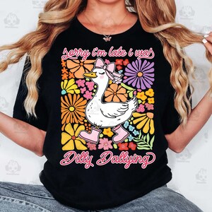 Op de afbeelding: Zwart T-shirt met een witte eend die roze laarzen en een roze strik draagt. De eend is omringd door kleurrijke bloemen. De tekst "Sorry I'm late I was dilly dallying" staat op het T-shirt gedrukt.