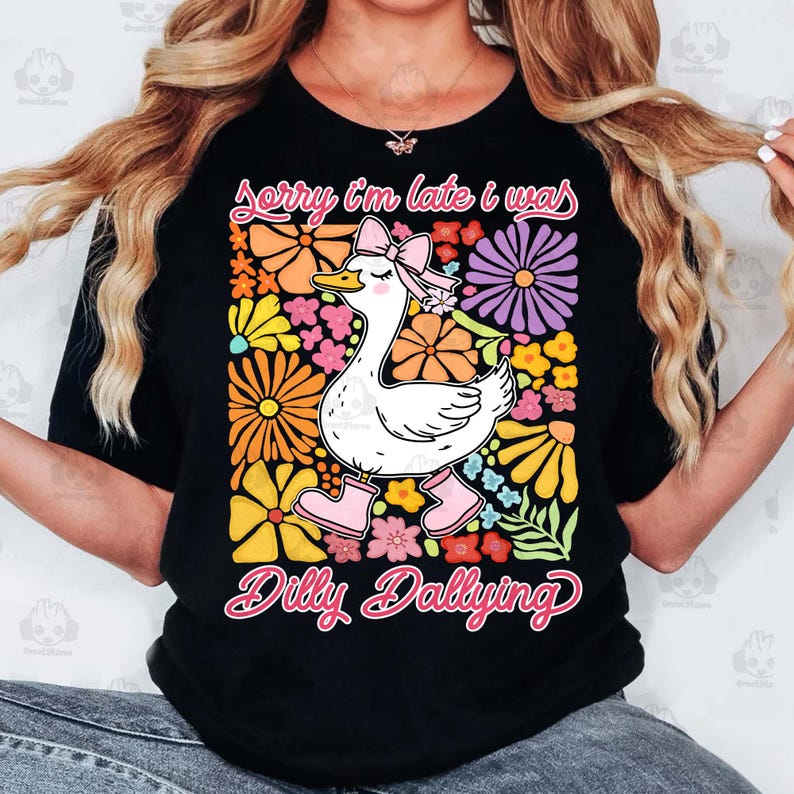 Op de afbeelding: Zwart T-shirt met een witte eend die roze laarzen en een roze strik draagt. De eend is omringd door kleurrijke bloemen. De tekst "Sorry I'm late I was dilly dallying" staat op het T-shirt gedrukt.