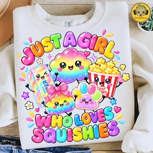 Może przedstawiać: Biała bluza z kolorowym nadrukiem graficznym z napisem JUST A GIRL WHO LOVES SQUISHIES. Wzór zawiera tęczową chmurę, kostkę masła, kubełek popcornu, kształt z kokardką oraz odcisk łapy, wszystkie z naniesionymi na nie uśmiechniętymi buźkami.