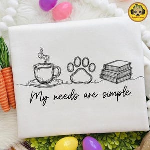 May include: White decorative towel with a black line drawing of a coffee cup, paw print, and books. The text "My needs are simple" is below the drawings. Easter eggs and carrots are in the background.