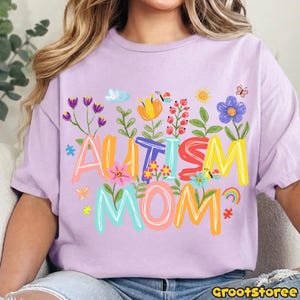 Puede incluir: Camiseta lila claro con las palabras "AUTISM MOM" en letras coloridas y florales. El dise&ntilde;o incluye flores, mariposas, un arco&iacute;ris y una pieza de rompecabezas.