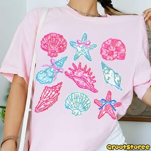 May include: Light pink t-shirt featuring a collection of colorful seashell and starfish illustrations. The designs include pink, blue, and white accents, with some adorned with bows. The shirt has a round neckline and short sleeves.