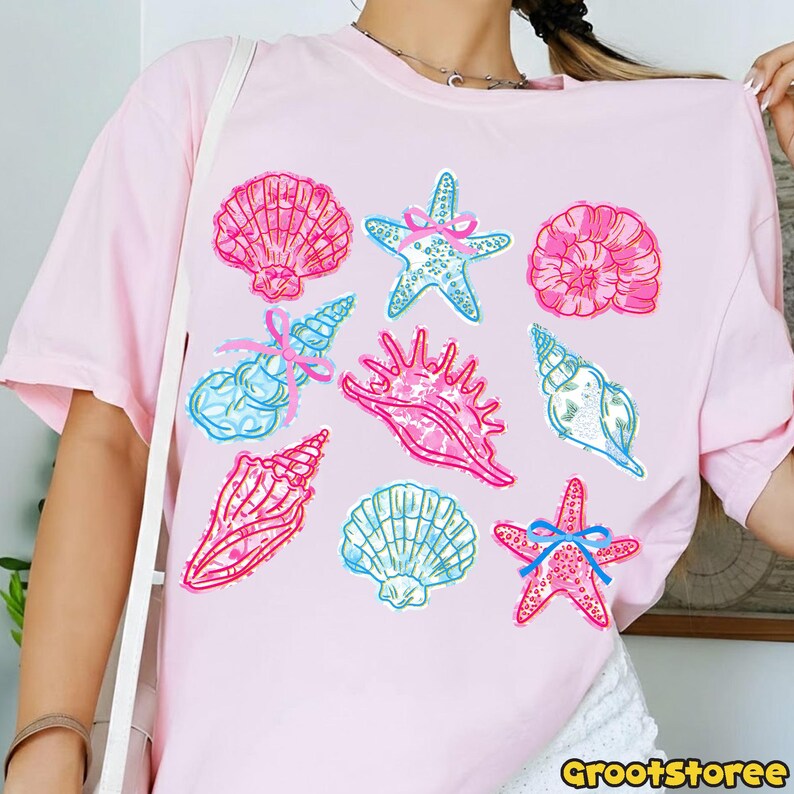 May include: Light pink t-shirt featuring a collection of colorful seashell and starfish illustrations. The designs include pink, blue, and white accents, with some adorned with bows. The shirt has a round neckline and short sleeves.