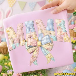 Puede incluir: Un bolso tote rosa claro con la palabra "MAMA" aplicada en un dise&ntilde;o de patchwork. Las letras presentan patrones florales y de cuadros, con un gran lazo en la parte inferior. El bolso es sostenido por una persona.