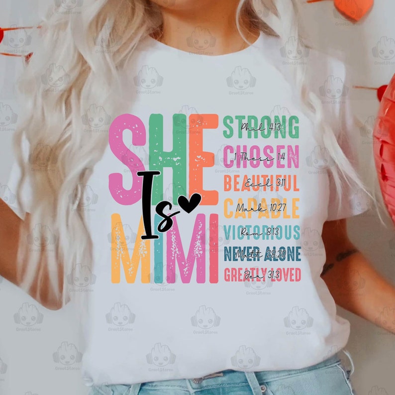 K&ouml;nnte beinhalten: Ein wei&szlig;es T-Shirt mit einer bunten Grafik, auf der "She is Mimi" steht, sowie Bibelverse und W&ouml;rter wie "Stark", "Auserw&auml;hlt", "Sch&ouml;n", "F&auml;hig", "Siegreich", "Niemals allein" und "Sehr geliebt".