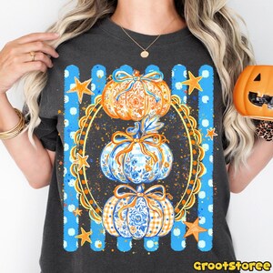 Puede incluir: Una camiseta gris carb&oacute;n con un gr&aacute;fico de tres calabazas apiladas dentro de un marco ovalado dorado ornamentado. Las calabazas tienen patrones florales y de cuadros en azul y naranja con lazos. El fondo tiene rayas verticales azules y estrellas.