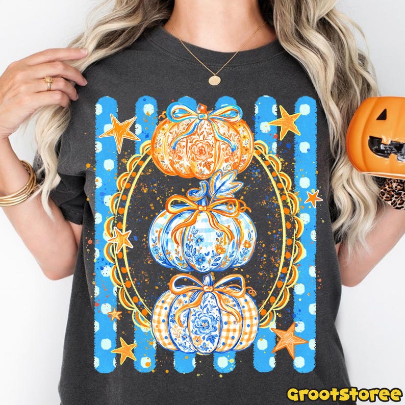 Puede incluir: Una camiseta gris carb&oacute;n con un gr&aacute;fico de tres calabazas apiladas dentro de un marco ovalado dorado ornamentado. Las calabazas tienen patrones florales y de cuadros en azul y naranja con lazos. El fondo tiene rayas verticales azules y estrellas.