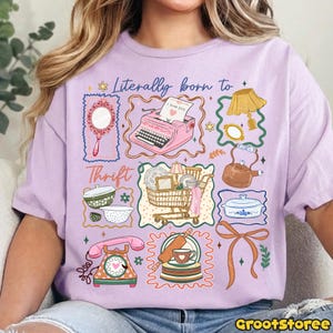 May include: A lilac-colored t-shirt featuring a whimsical graphic design. The design includes a pink typewriter, a shopping cart, a telephone, and other vintage-themed illustrations. The text "Literally born to Thrift" is also present.