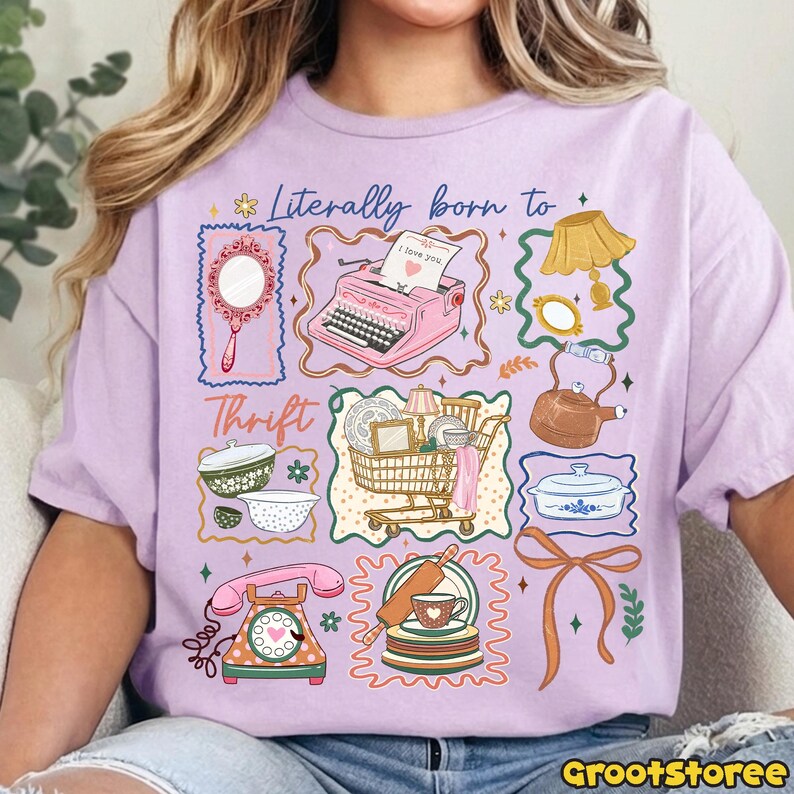May include: A lilac-colored t-shirt featuring a whimsical graphic design. The design includes a pink typewriter, a shopping cart, a telephone, and other vintage-themed illustrations. The text "Literally born to Thrift" is also present.