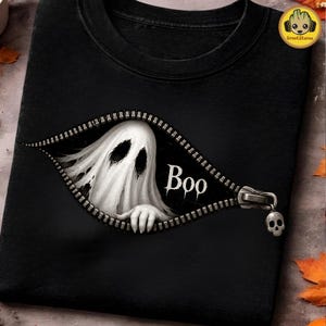 May include: A black t-shirt featuring a graphic of a white ghost peeking through an open zipper. The ghost has dark, hollow eyes and a hand reaching out. The word "Boo" is printed in white next to the ghost, and the zipper pull is a skull.