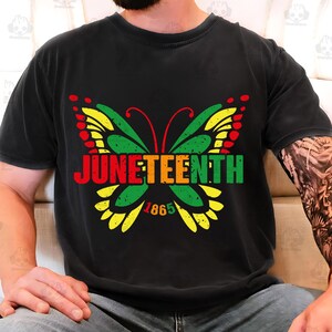 May include: Black t-shirt featuring a colorful butterfly design with the word "Juneteenth" in red, orange, green, and yellow. The year "1865" is below the butterfly. The shirt is a comfortable, casual style.
