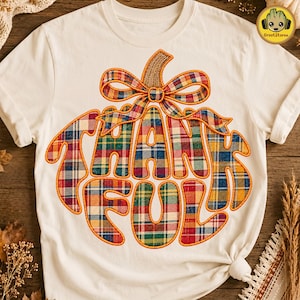 Op de afbeelding: Een crèmekleurig t-shirt met een grote afbeelding in de vorm van een pompoen, gevuld met het woord THANKFUL in een veelkleurig ruitpatroon. De pompoen is bekroond met een strik in ruitpatroon en heeft een gestikt omtrekeffect.