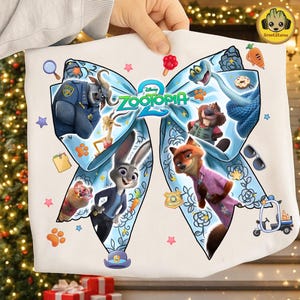 May include: White Zootopia-themed graphic tee featuring characters like Judy Hopps and Nick Wilde. The design is centered around a large blue bow with floral accents and the movie's logo. Additional elements include a police badge, carrot, and paw prints.