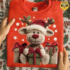 Könnte beinhalten: Rotes Sweatshirt mit einem Plüsch-Rentier-Design mit Weihnachtsmütze, Geweih und roter Nase. Das Rentier hält drei verpackte Geschenke mit roten und grünen Schleifen. Weiße Schneeflocken sind verstreut.
