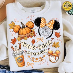 May include: A white crewneck sweatshirt featuring a colorful autumn-themed graphic. The design includes orange pumpkins, a coffee cup with whipped cream, a spiderweb-patterned headband with ears, and the text 'TIS THE SEASON' on a banner.