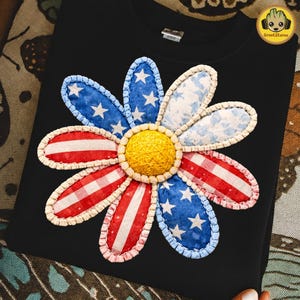 May include: Black sweatshirt featuring a large, appliqued flower design. The flower petals incorporate patriotic patterns: red and white stripes, blue with white stars, and a light blue floral print. The center is a yellow, textured sphere.
