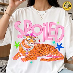 May include: White t-shirt with a graphic of a leopard wearing a crown and necklace, with the word "SPOILED" in pink. The leopard is orange with black spots, and the design includes colorful star accents.