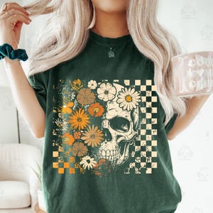 May include: Dark green t-shirt featuring a skull design with floral and checkered elements. The skull is adorned with daisies and other flowers in shades of orange, white, and brown. A checkered pattern is on the right side of the skull.
