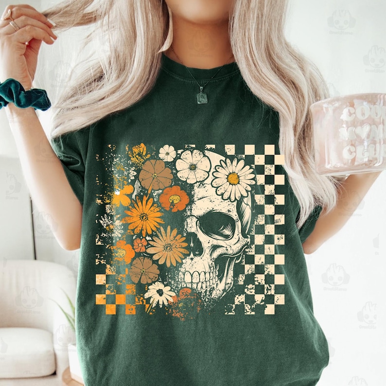 May include: Dark green t-shirt featuring a skull design with floral and checkered elements. The skull is adorned with daisies and other flowers in shades of orange, white, and brown. A checkered pattern is on the right side of the skull.