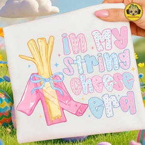 Puede incluir: Camiseta blanca con un gráfico caprichoso. El diseño presenta un manojo de queso en hebras envuelto en una chaqueta rosa y un lazo azul. El texto dice "In My String Cheese Era" en letras coloridas y estampadas. Huevos de Pascua y hierba en el fondo.