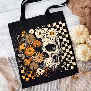 May include: Black tote bag featuring a skull design with floral accents in shades of orange, brown, and cream. The design includes a checkered pattern and a grunge aesthetic. The bag has black handles.