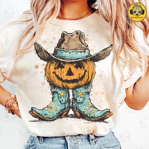 Può includere: T-shirt color crema con grafica a tema Halloween. Il design raffigura una zucca intagliata con cappello da cowboy e stivali da cowboy turchesi. La zucca ha una faccia sorridente. La grafica è nei toni dell'arancione, del marrone e del blu.
