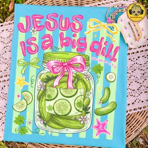 May include: A light blue t-shirt featuring a graphic of a glass jar filled with green pickles, tied with a pink ribbon. The text reads JESUS IS A BIG DILL and Philippians 2:9-10. The design includes yellow bows, stars, and small flowers.