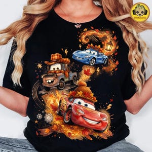 May include: A black t-shirt featuring a graphic print of three animated cars. A brown tow truck wears a plaid hat and scarf, a blue sports car is near a lantern, and a red race car wears a knit hat, all surrounded by autumn leaves.